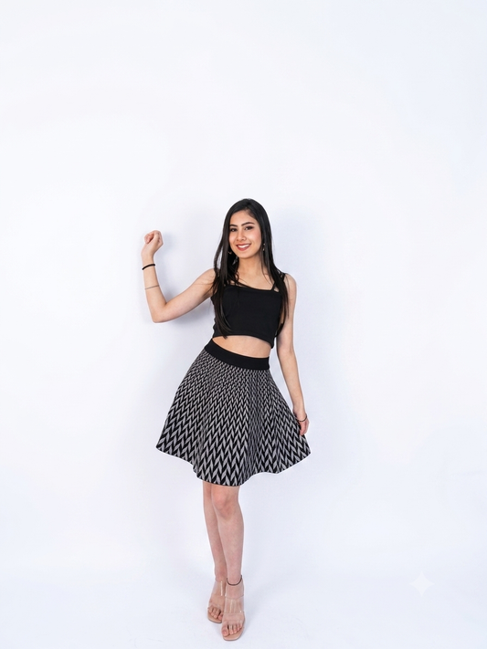 A woman modeling a black sleeveless crop top and a black and white patterned pleated mini skirt, standing against a solid white studio background.